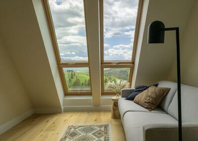 Pokój z ukośną ścianą i oknami z widokiem. Kanapa z lampką nocną w szarym kolorze.