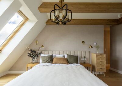 Sypialnia w naturalnych kolorach z podwójnym łóżkiem. Ściana pod skosem z dużym oknem. Meble z naturalnego drewna.