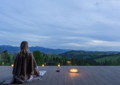 kobieta siedząca na tarasie z widokiem na góry, medytująca. Na tarasie płonie palenisko. Panuje wieczór