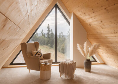 pokój na poddaszu z drewnianym, jasnym sufitem, wygodnym białym fotelem, pufą oraz dekoracyjnym wazonem.